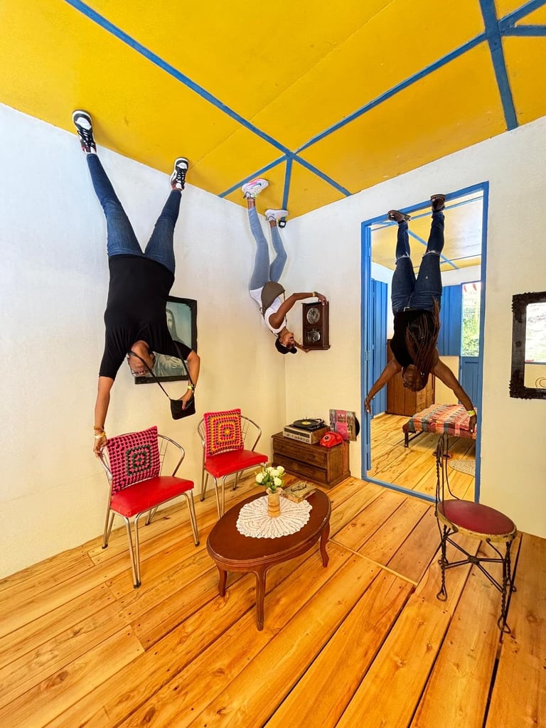 Two people performing aerial yoga inversions upside down in a bright room with yellow ceiling, wooden floor, and red chairs