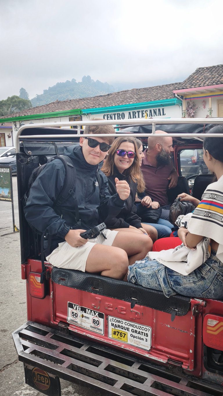 Group of five people sitting in the back of a red pickup truck with mountain buildings visible in background